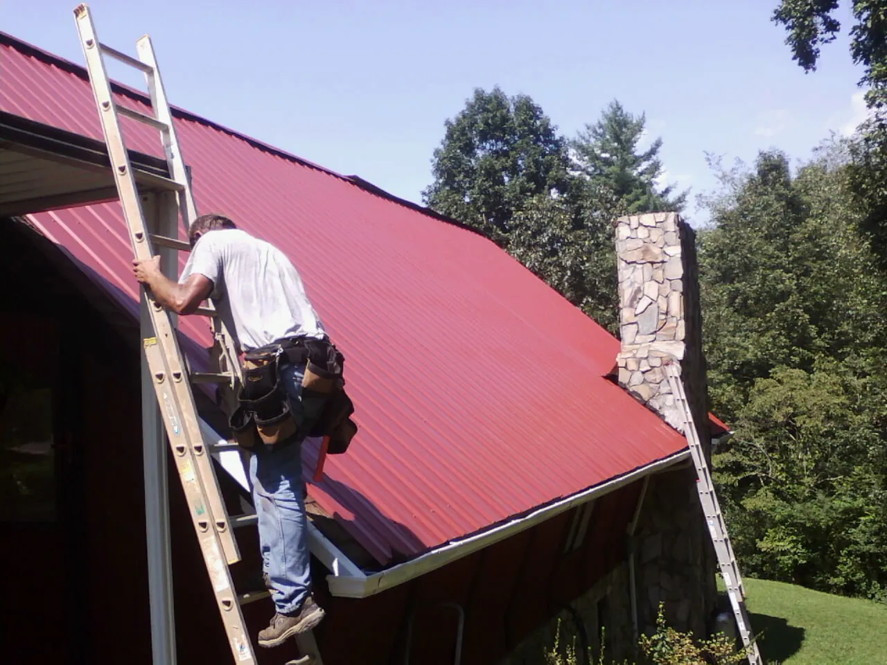 Roofer on a ladder beside a red metal roof and stone chimney — Soffit & Fascia Repair by Roofing Brothers in Cabins